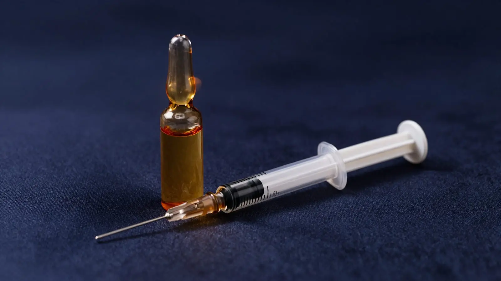 Amber-gold glass vial of peptide serum and a fine syringe resting on deep navy silk